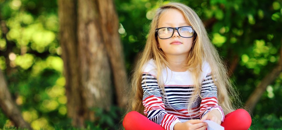 Young child wearing glasses sitting cross-legged in front of a tree with a book in front of her looking at the camera
