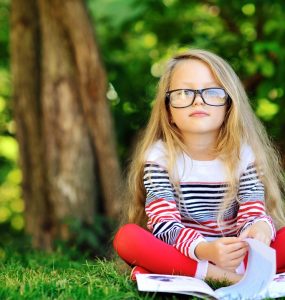 Young child wearing glasses sitting cross-legged in front of a tree with a book in front of her looking at the camera