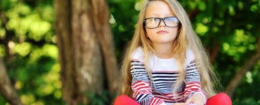 Young child wearing glasses sitting cross-legged in front of a tree with a book in front of her looking at the camera