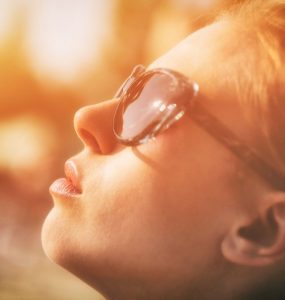 Close up photo of a woman wearing sunglasses looking up at the sun being bathed in a yellow glow