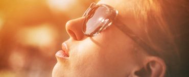 Close up photo of a woman wearing sunglasses looking up at the sun being bathed in a yellow glow