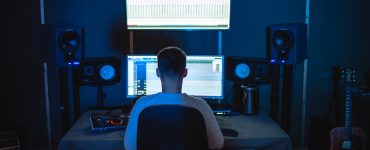 Young man sitting in a dark room filled with music equipment in a chair in front of an ultrawide computer screen.
